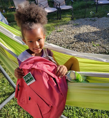 Photo of smiling young girl in a hammock holding a backpack.